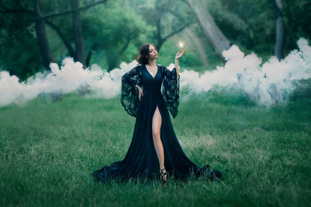 woman in a black long dress with a trailer and lace sleeves. magic. witchcraft. leg, slender figure.stealing soul, spirit hunter, fire in hands, sparks, mysteries, smoke, forest, art photo. sun light.
