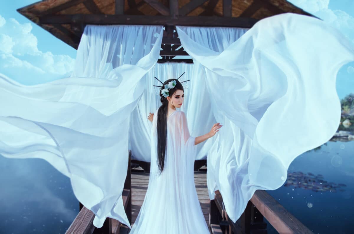 A beautiful sorceress stands on the background of a wooden gazebo by the water, with a white air dress. In the picture there are many flying curtains and silk fabrics. Japanese hairstyle
