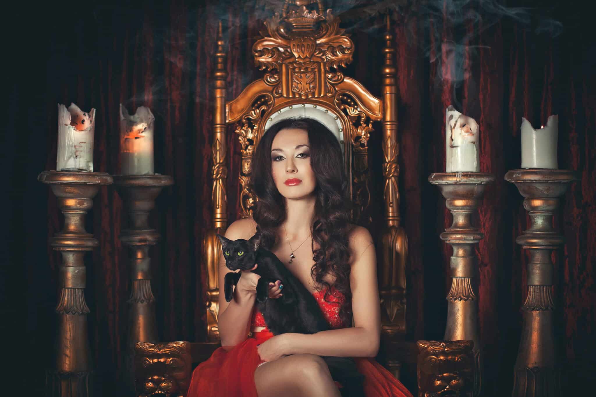 A brunette girl in a red dress sitting on a throne with a black cat in her arms against a background of candelabra with candles.
