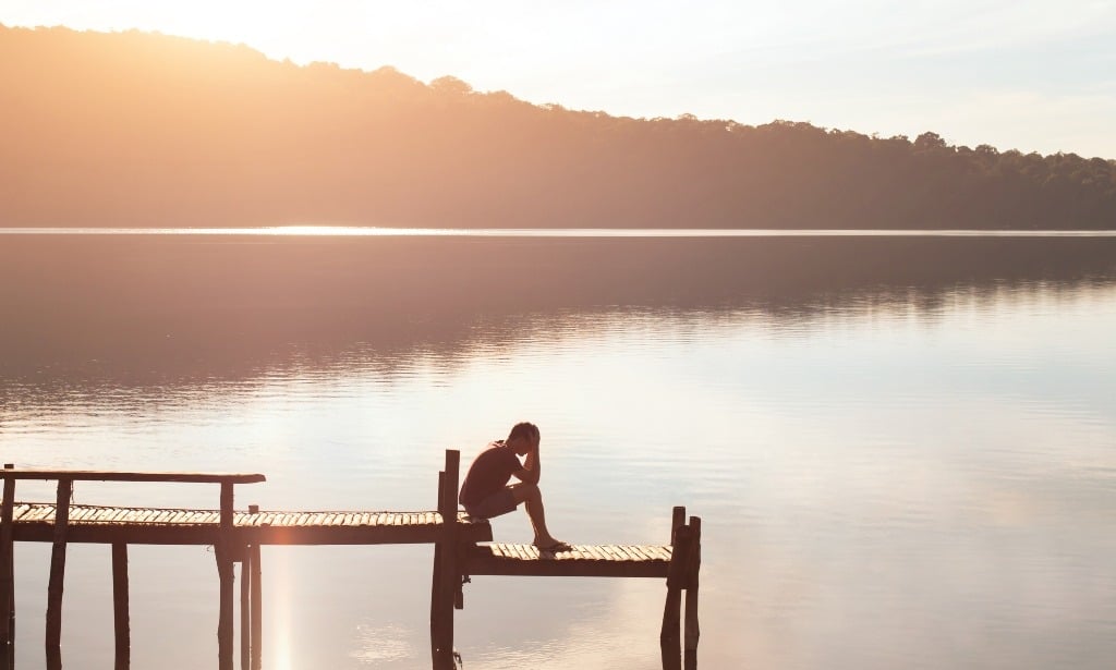 Depressed man sitting on a wooden dock at sunrise.