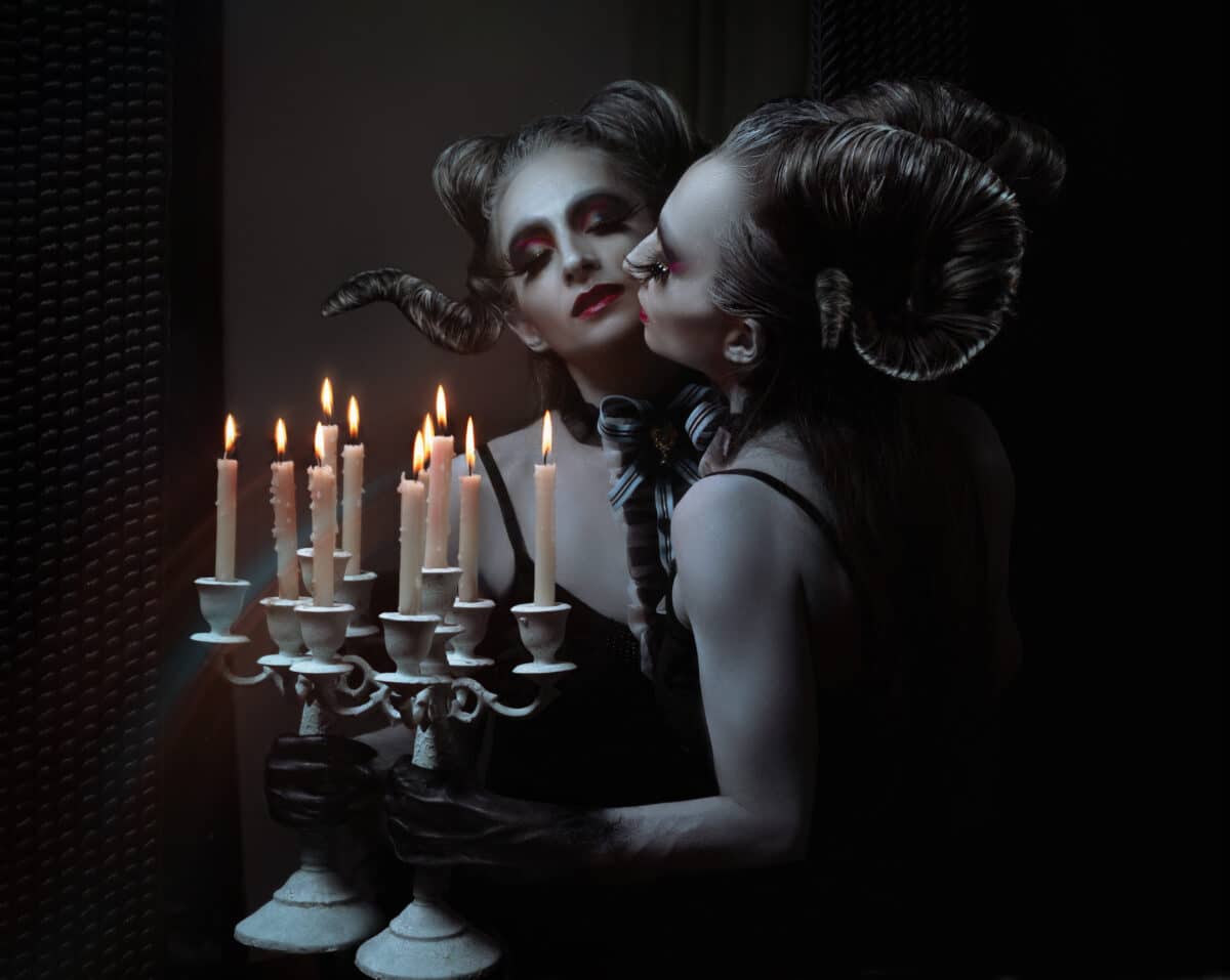 Portrait of beautiful Halloween monster woman with horns looking at mirror and holding candlestick in darkness