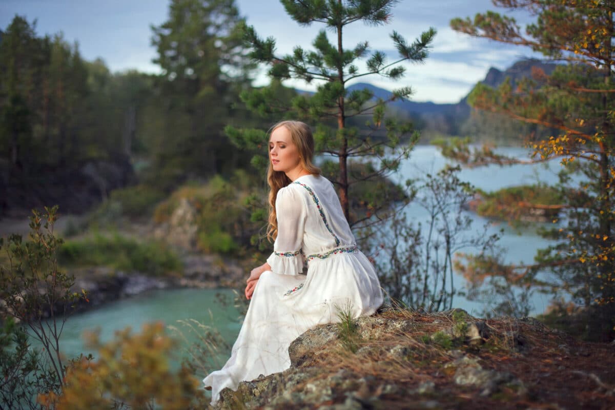 A girl in a white dress walks on a rocky shore near the river.