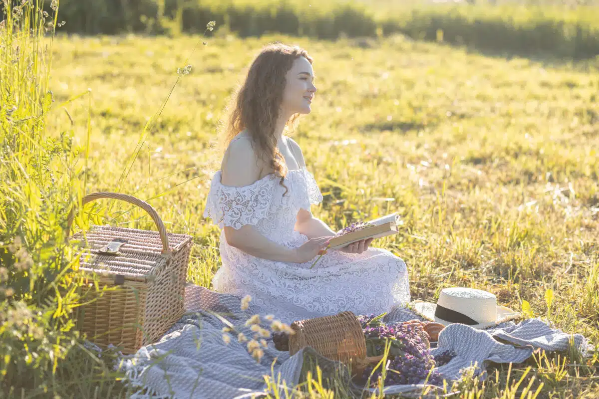 Beautiful young girl in a white dress, straw hat, picnic basket reading a book on a meadow. Summertime, golden hour, sunset. Work life balance concept