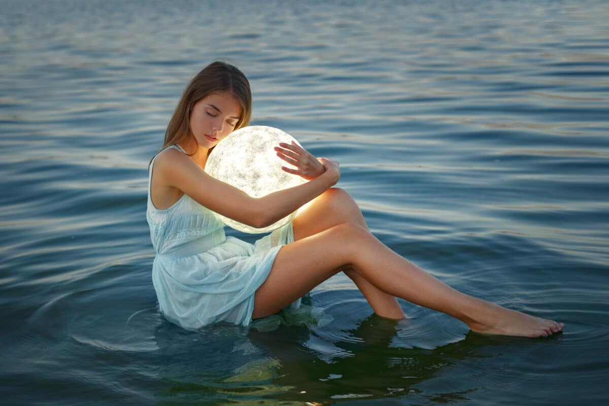 Attractive young girl in a blue dress sits on the water and holds the moon in her hands