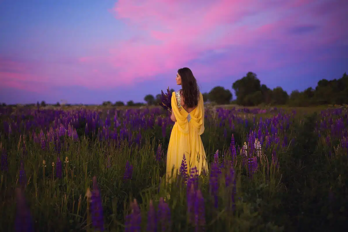 A beautiful girl in a long yellow dress against the background of a blooming purple field and a bright sunset sky