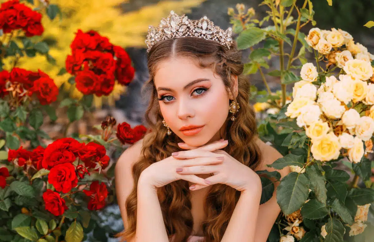 Fantasy girl princess beautiful face, delicate make-up, a crown of jewels on head, long hair. Happy woman sits in garden enjoys nature, green bushes red flowers roses. art pink white vintage dress