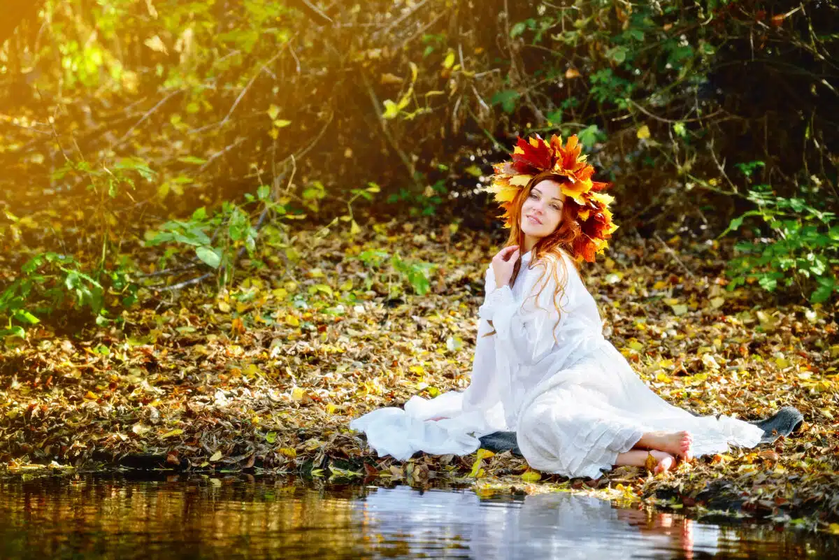 woman in a white dress wearing a head wreath is sitting on the river bank 
