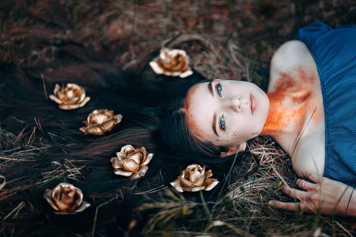 portrait of beautiful brown-haired woman in a blue dress lying on grass, with golden roses on her long hair, in the fairy forest, golden paint shimmer on her neck fantasy concept