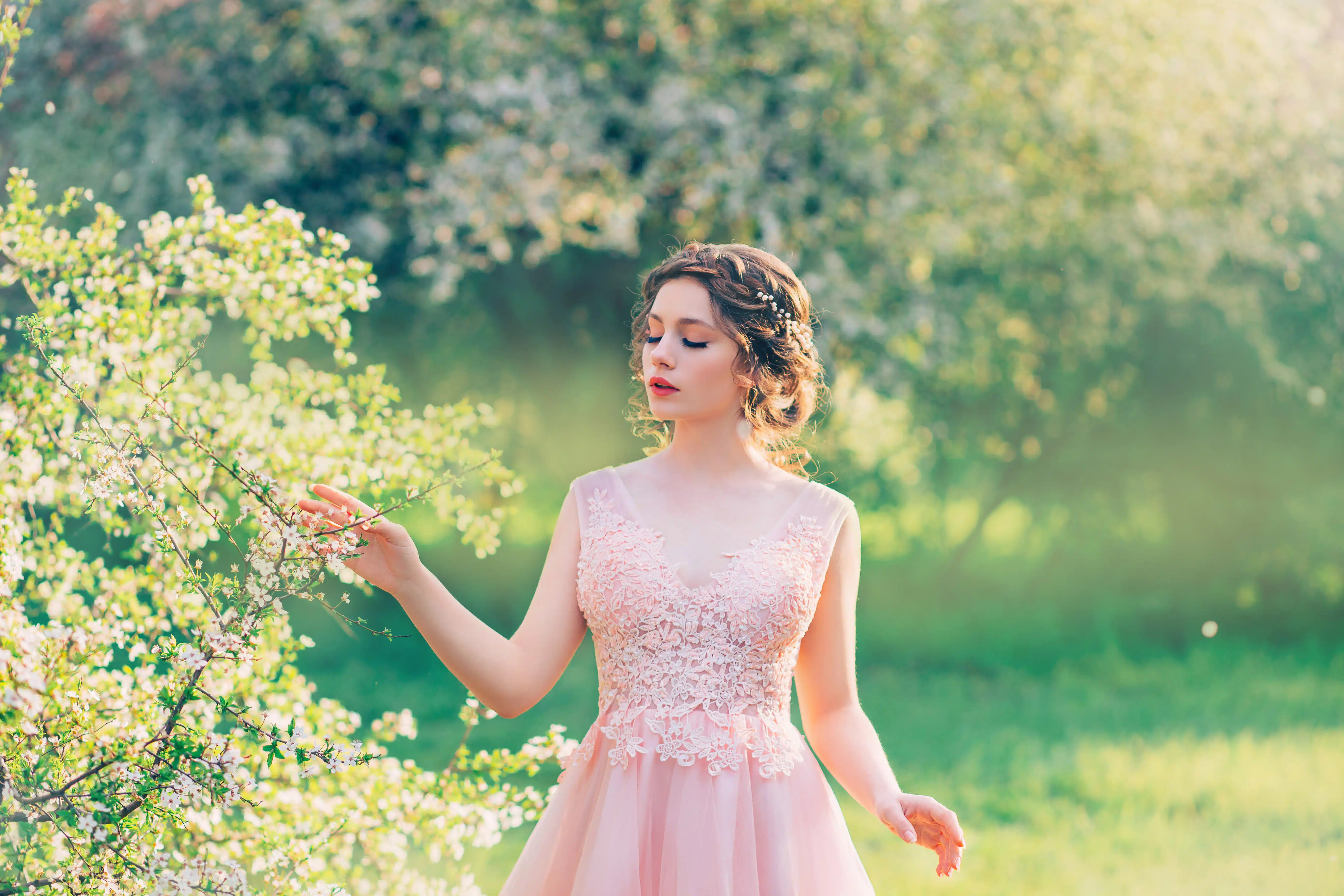 charming lady in blooming garden, girl with gathered hair gently strokes branches of trees with flowers, porcelain doll with bright red cute lips in pink dress, image of spring nymph, creative colors