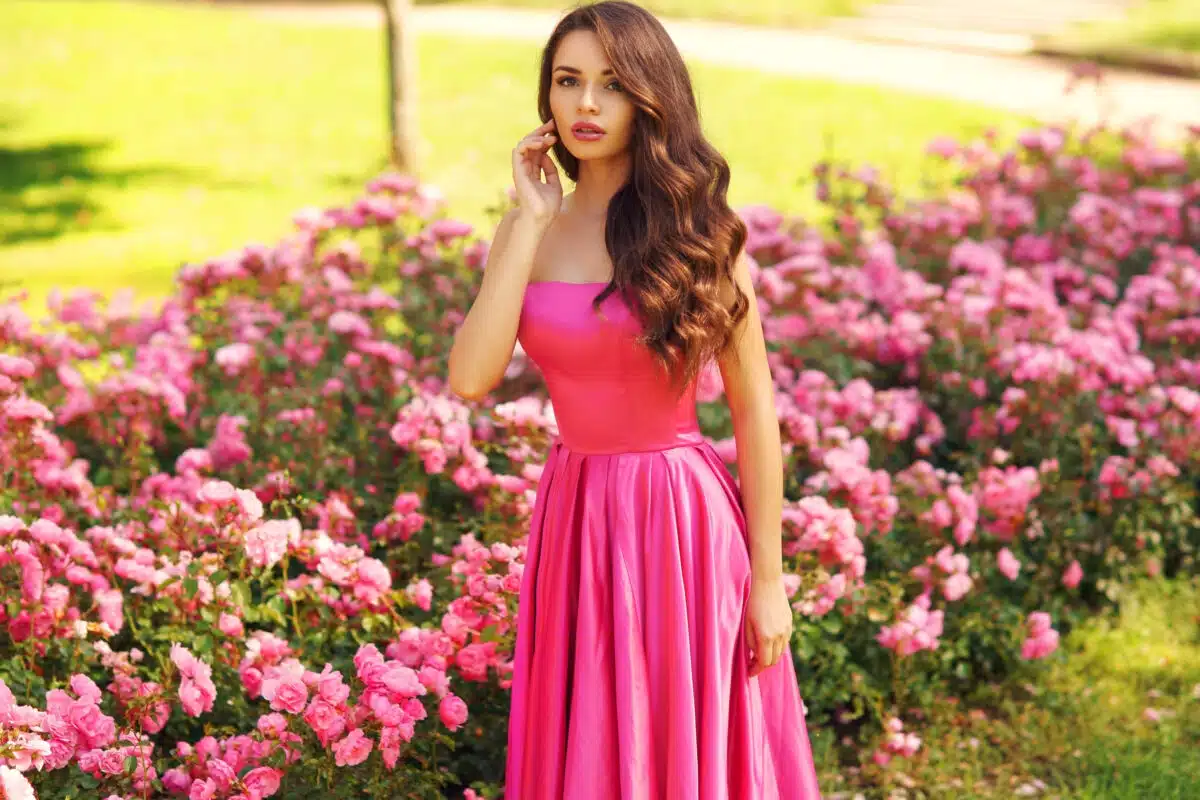 Young beautiful pretty woman posing in long evening luxury dress against bushes with pink roses on a sunny summer day