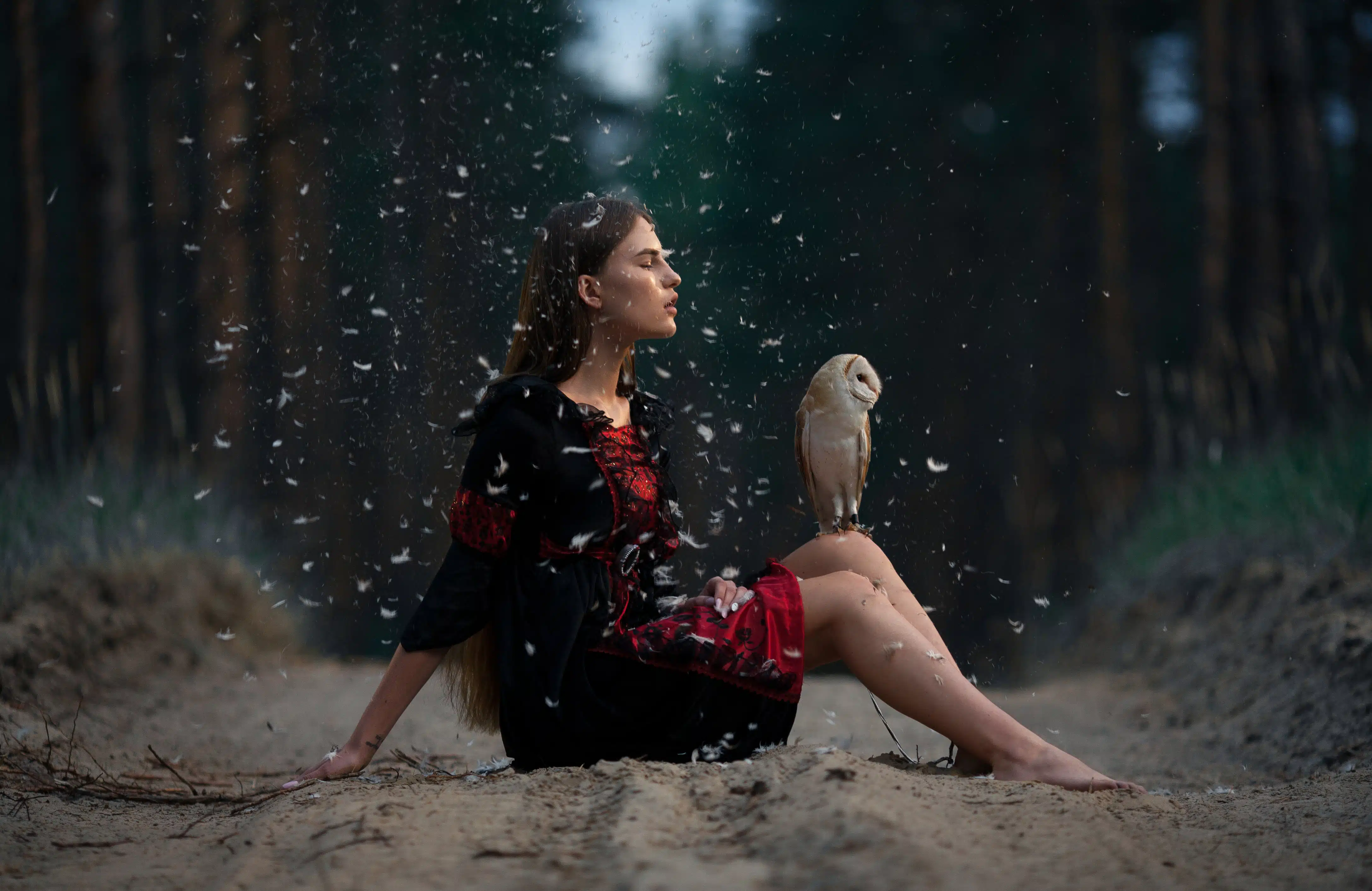 young woman sits on forest road with owl on her knees among flying feathers