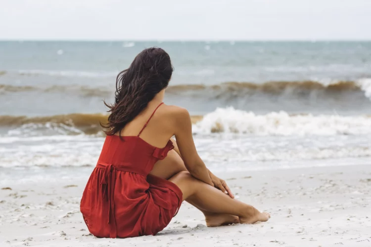 Woman in red summer dress sitting on the beach looking toward the wavy ocean