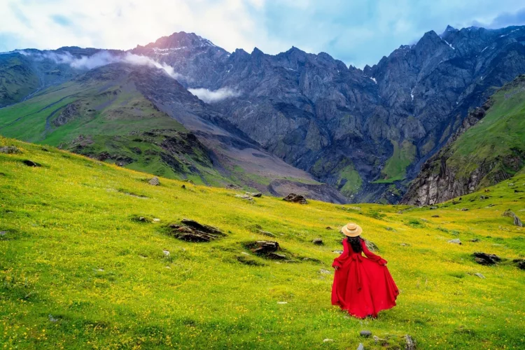 woman in red dress enjoy view of green pasture and flowers near the mountain