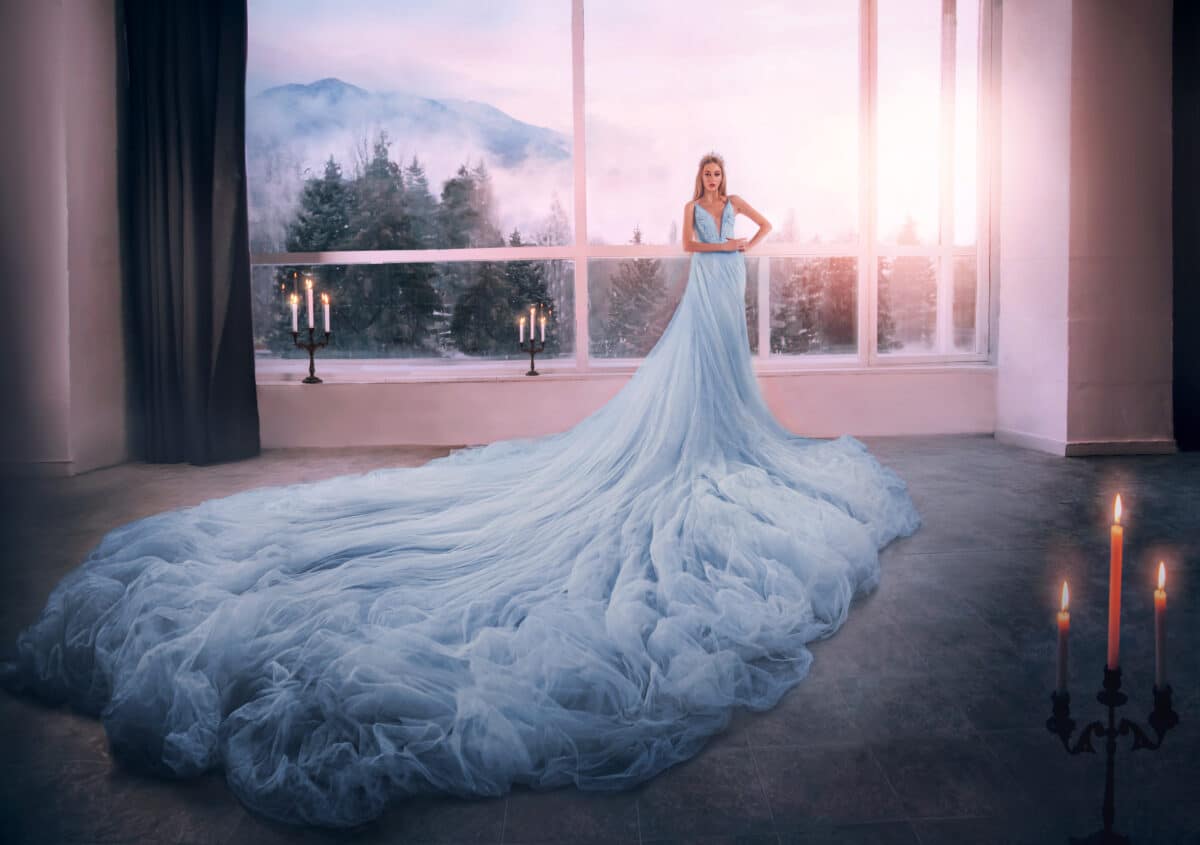 Young woman blonde fantasy princess Cinderella in glamorous blue dress stands in white room huge window. Long skirt tulle train hem. Romantic elegant image medieval queen, crown. Historical lady girl