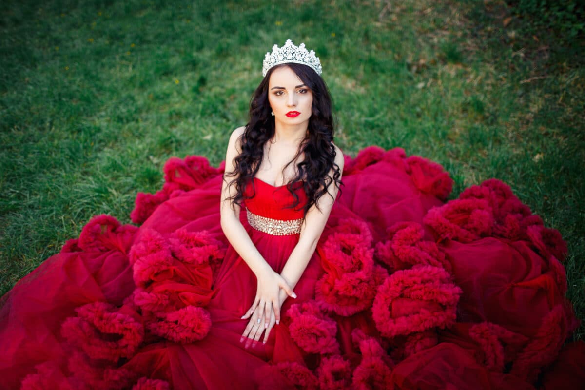 beautiful brunette in a crown with a bouquet in his hand and in a chic big and lush red dress in the park among the green