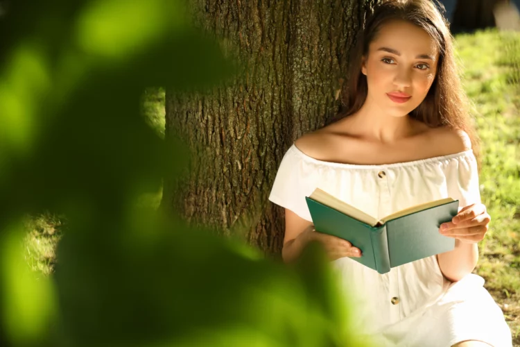 Beautiful young woman reading book near tree in park