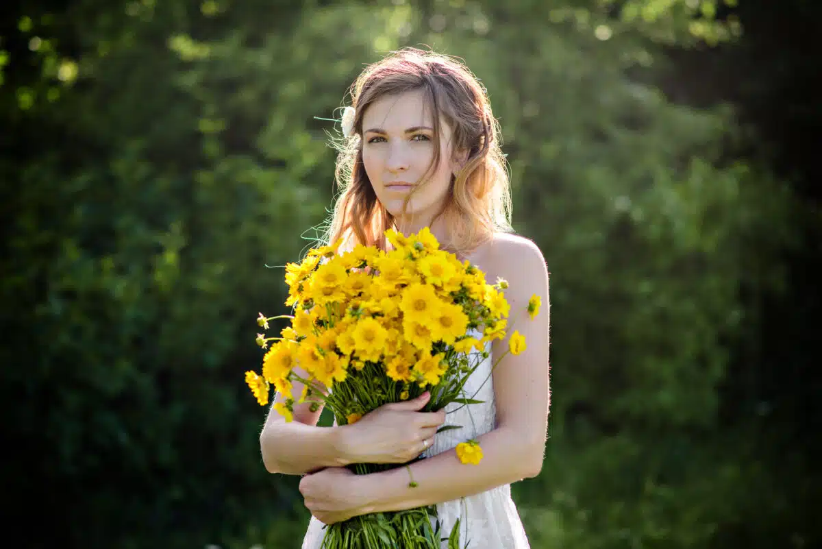 A lady in a white dress holds a huge bouquet of yellow flowers outdoor