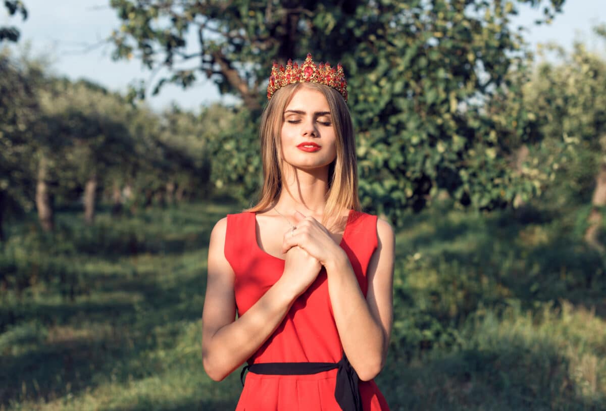 Blonde in bright red dress with a diadem on her head is standing outdoor in the garden