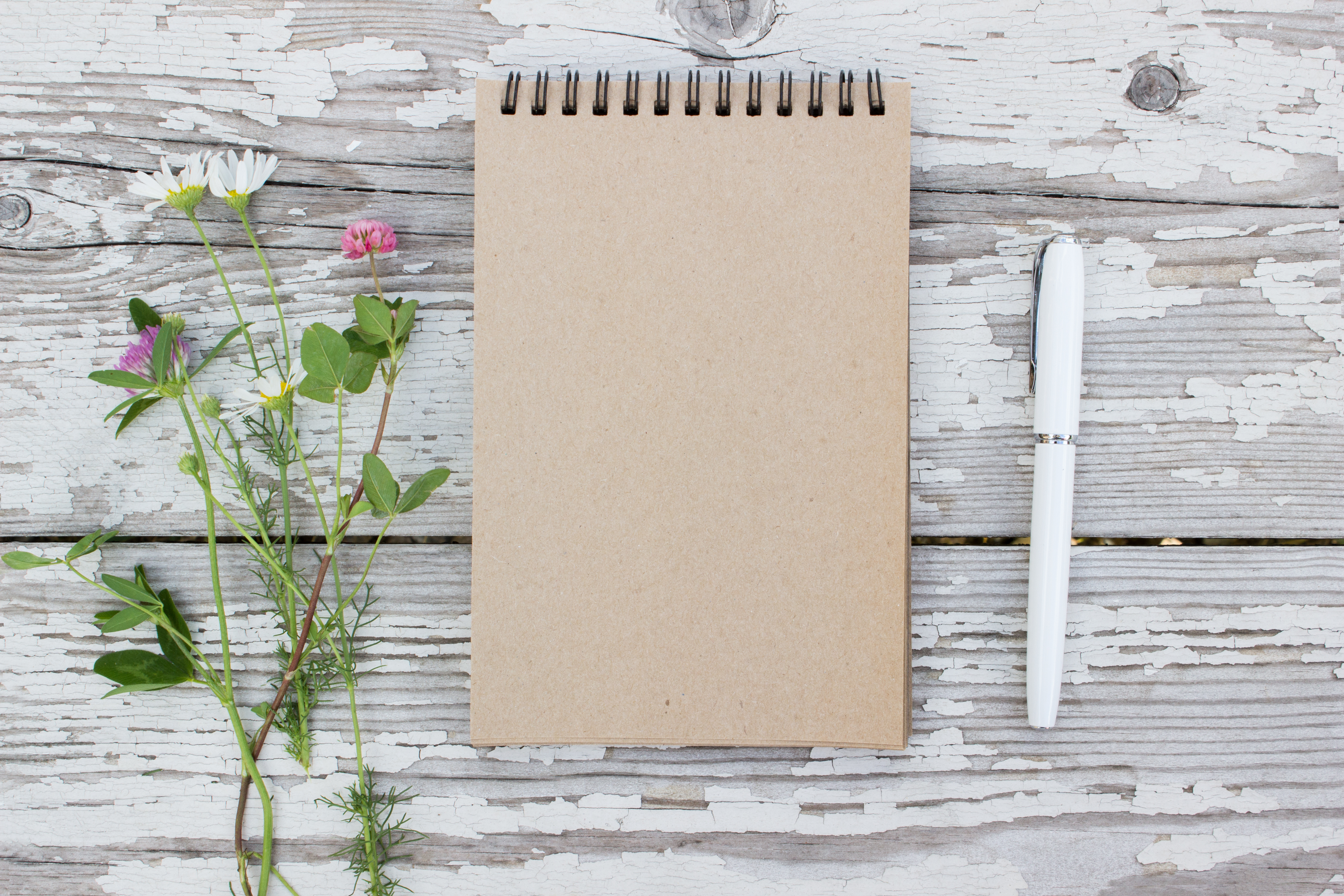 Kraft paper spiral notebook, white pen and summer flowers on rustic wooden table.