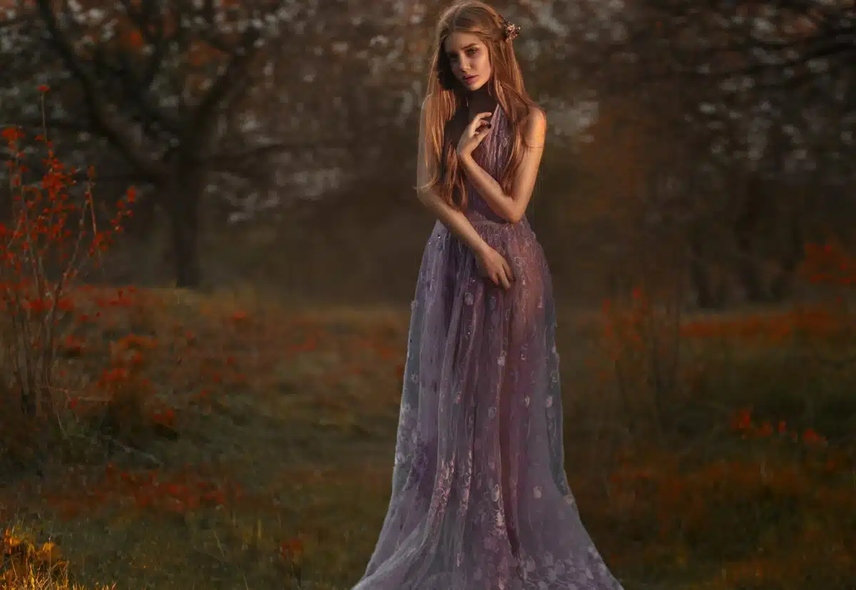 a sad blonde lady is standing in the garden at sunset in a luxurious purple dress