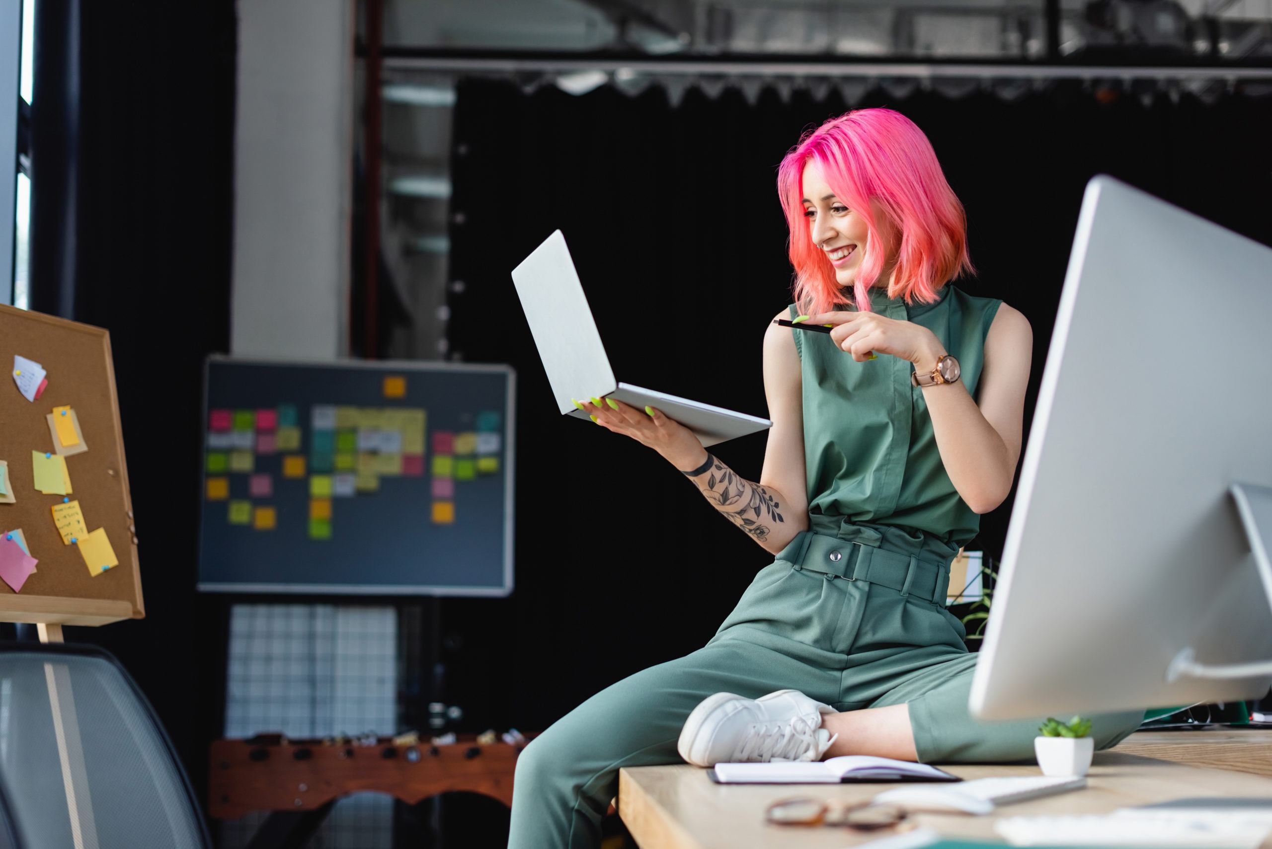 cheerful woman with pink hair pointing with pen at laptop near monitor in office.
