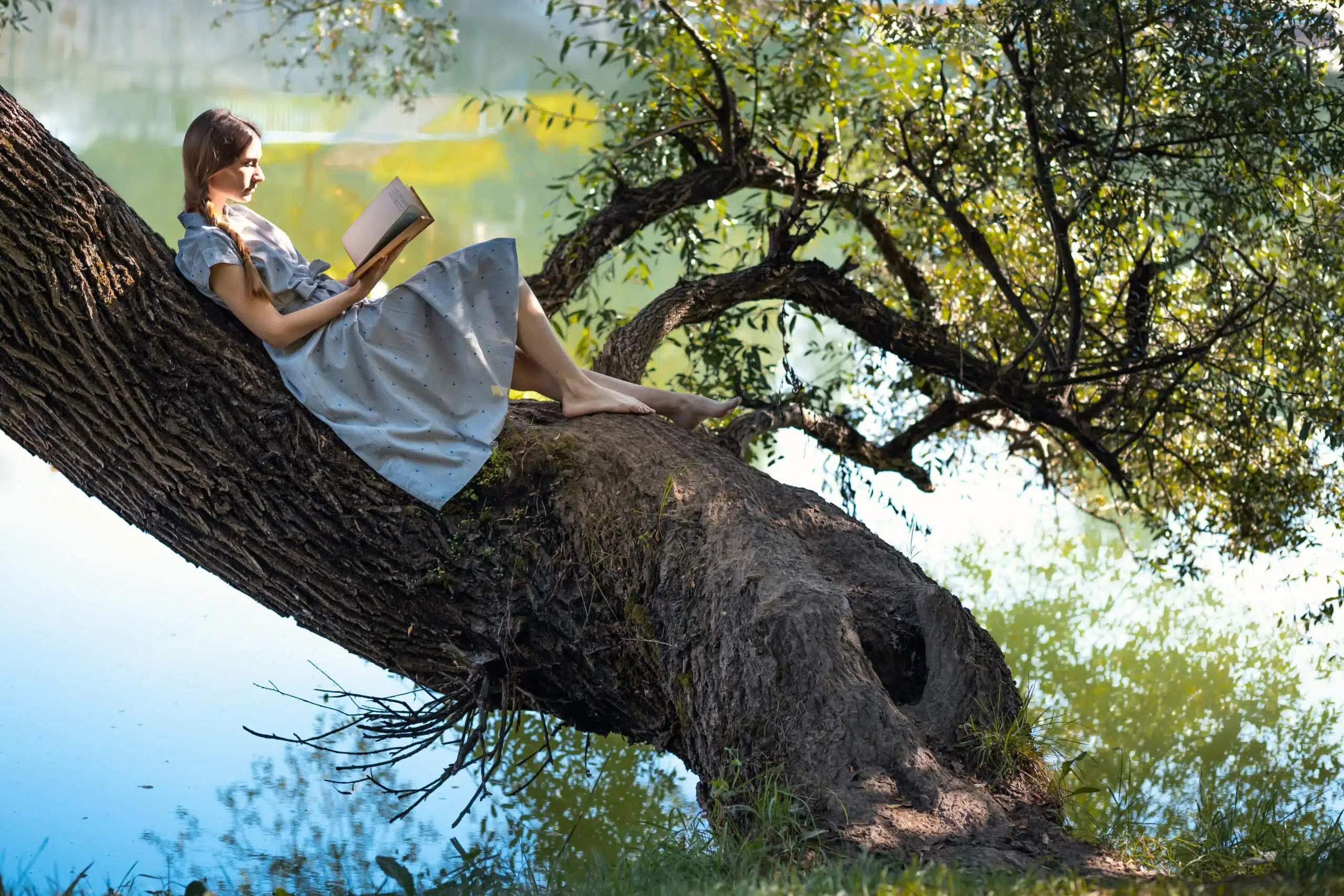 Young girl sitting on a tree by the river, reading a book.