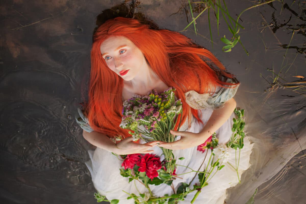 Redhaired girl in a romantic dress lies with open eyes in the wa