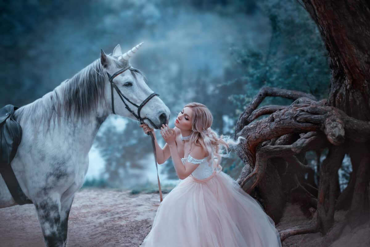 A fairy in a tender vintage dress hugs a unicorn. Fantastic magical, radiant horse. Background river and forest. Blonde girl with wavy hair - light elf.