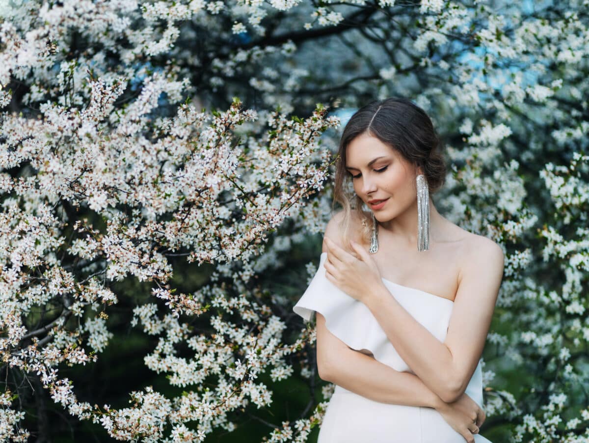 Portrait young beautiful woman fashion model posing. background blooming tree spring nature white flowers. Elegant girl romantic collected hairstyle. long trendy stylish earrings jewelry accessories