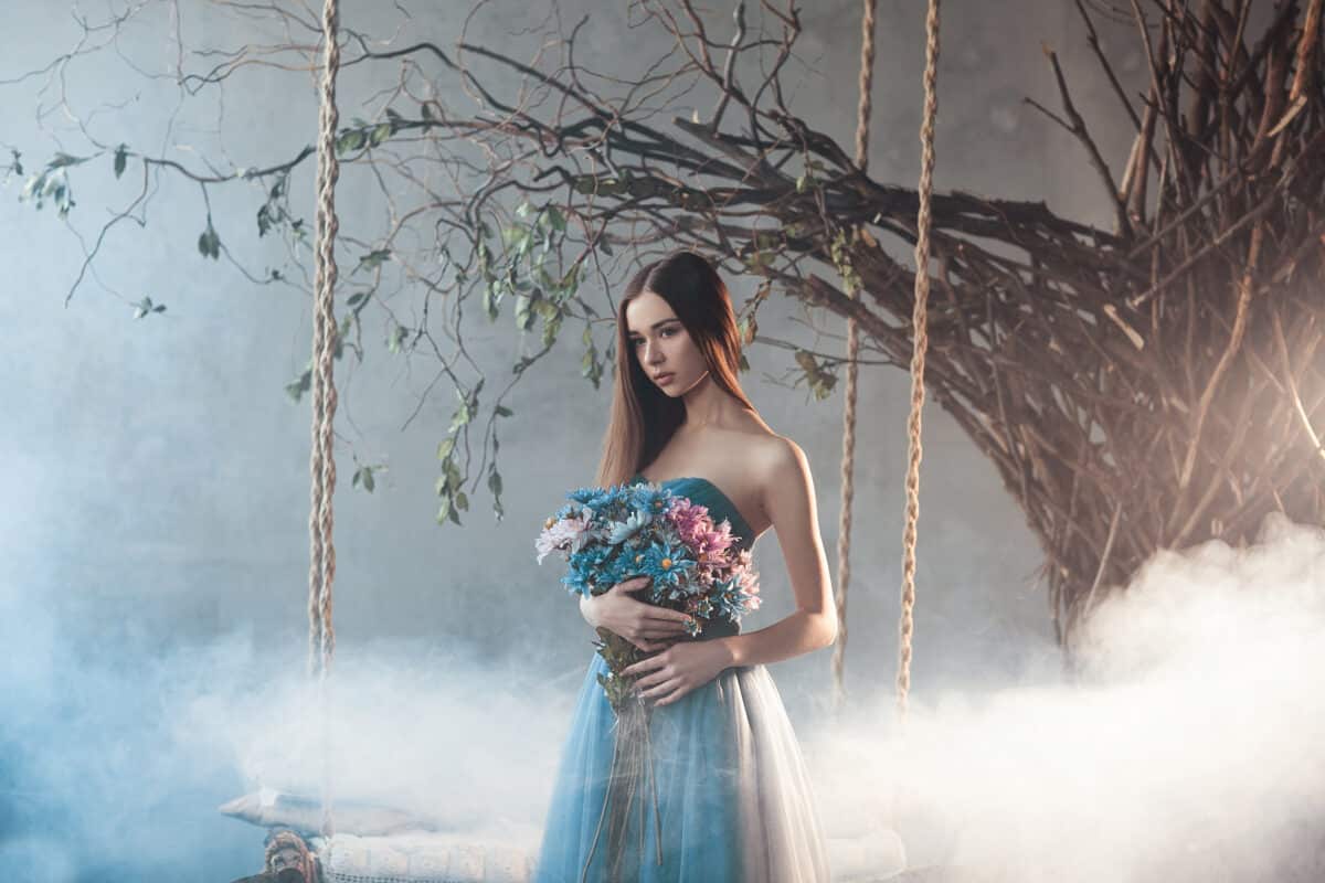 Beautiful gorgeous calm woman in long chiffon blue dress holding big bouquet of flowers standing in the forest near the tree with swing. Fantastic unusual atmosphere with fog in the forest. Wood Fairy