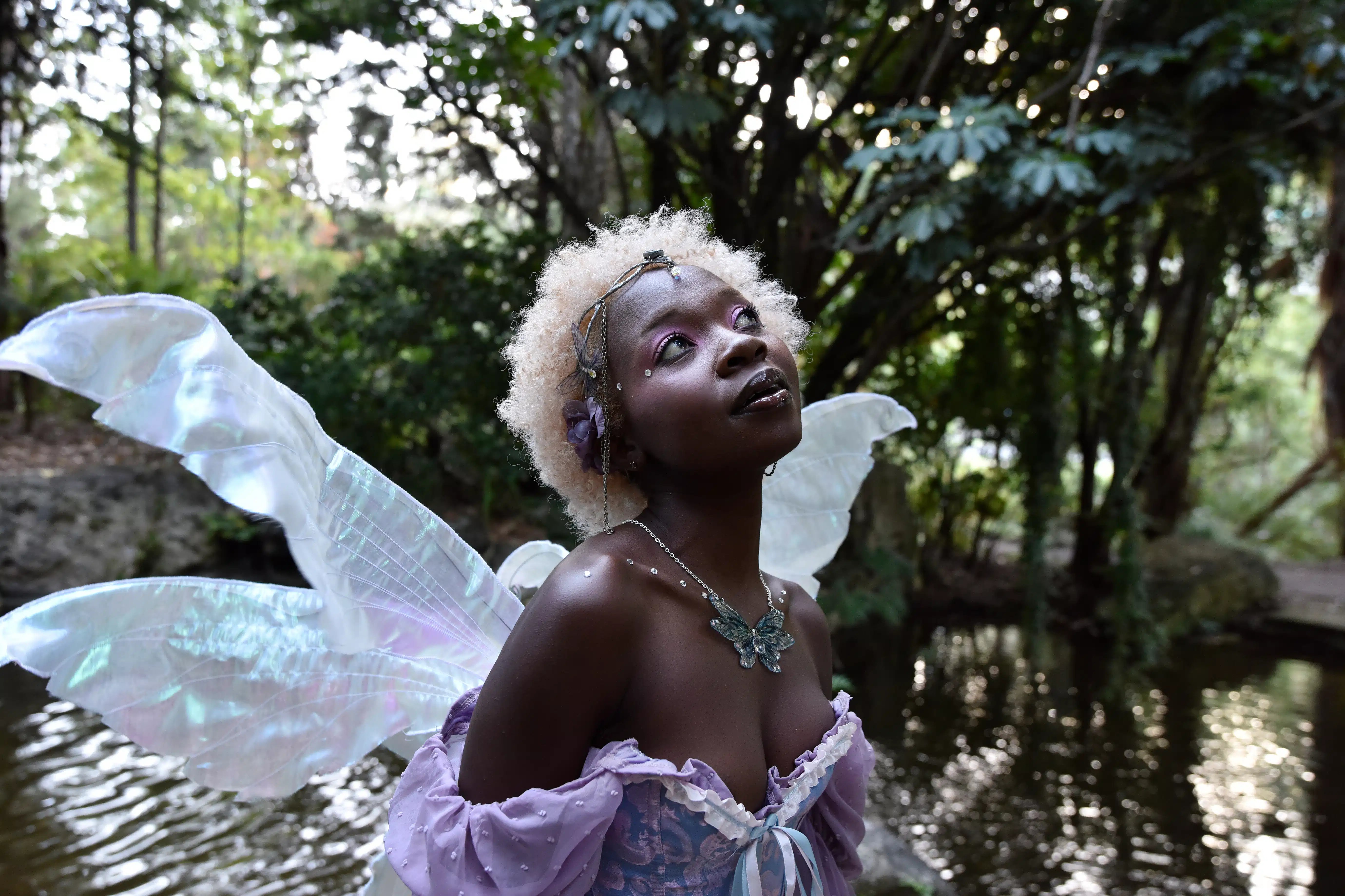 Portrait of beautiful African woman wearing purple fantasy costume, magical fairy wings and flower crown afro, wandering around forest location with natural lighting.