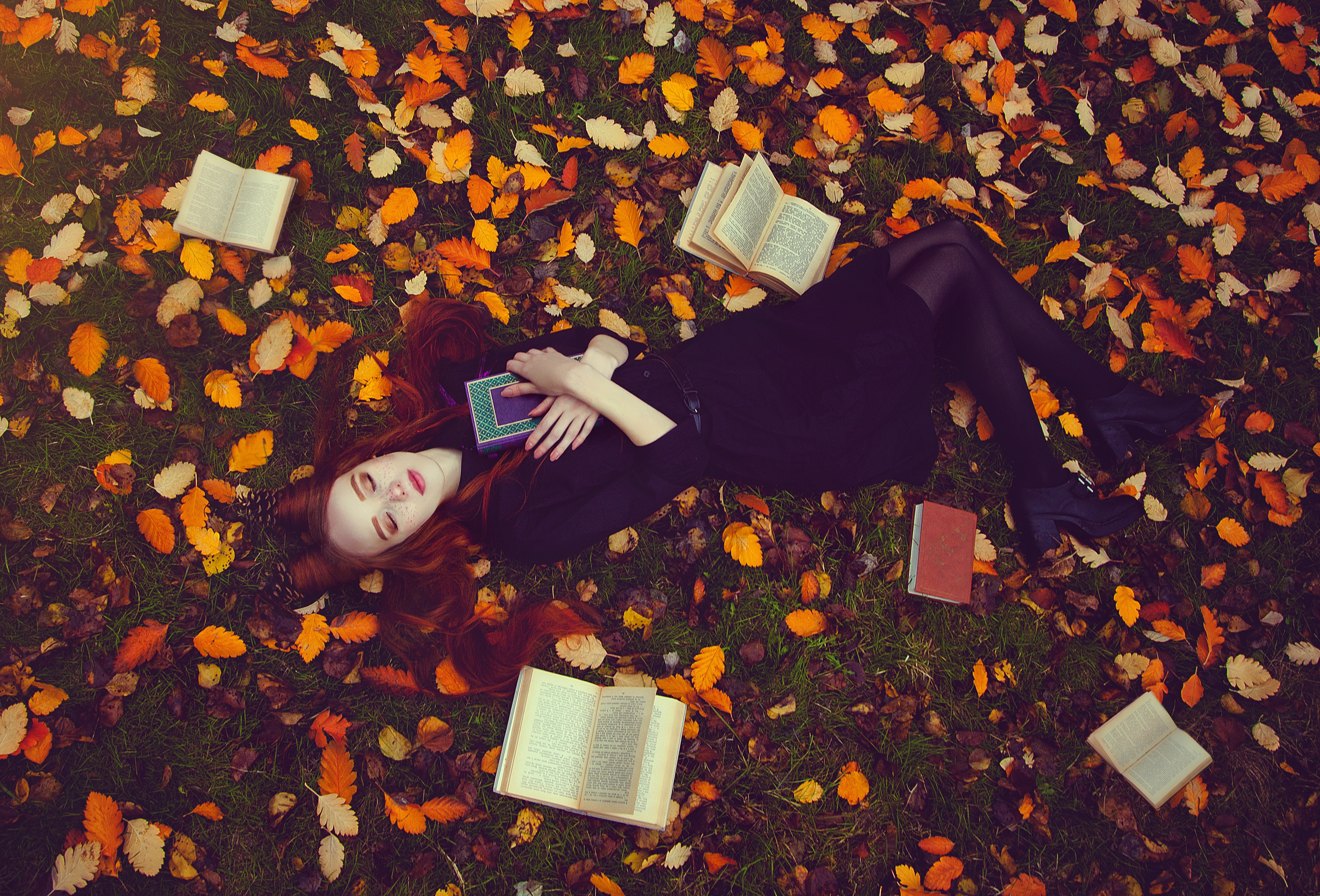 Beautiful red-haired girl with books lies on the grass in an autumn forest, top view. Autumn Fairytale photoshoot.
