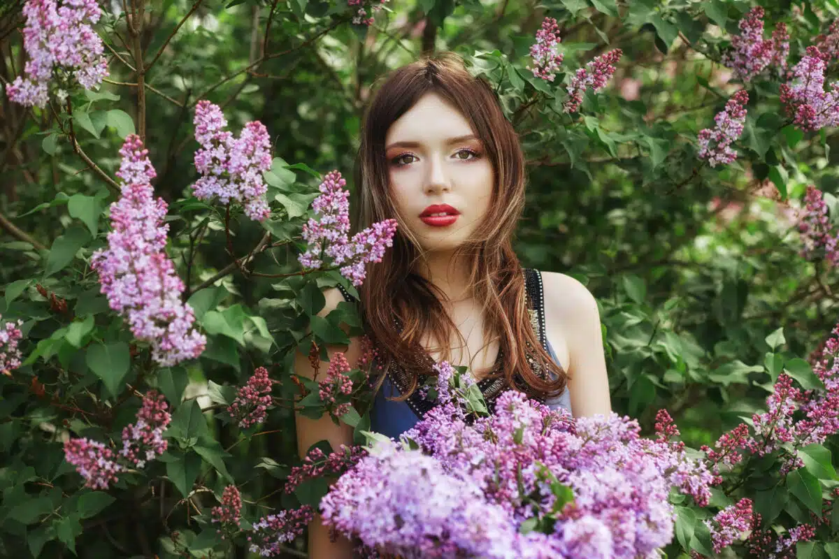 Beautiful girl in a dress posing near a Bush of lilacs on a summer day, purple flowers in the Park. Spring portrait of a girl in nature in the sun. Funny emotions on his face