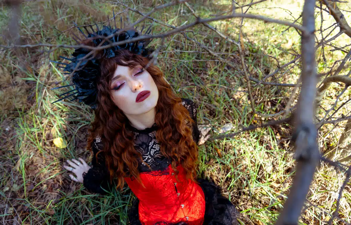 Top view through dry branches on a young woman in a carnival costume sits on the grass with her eyes closed. Halloween woman portrait.