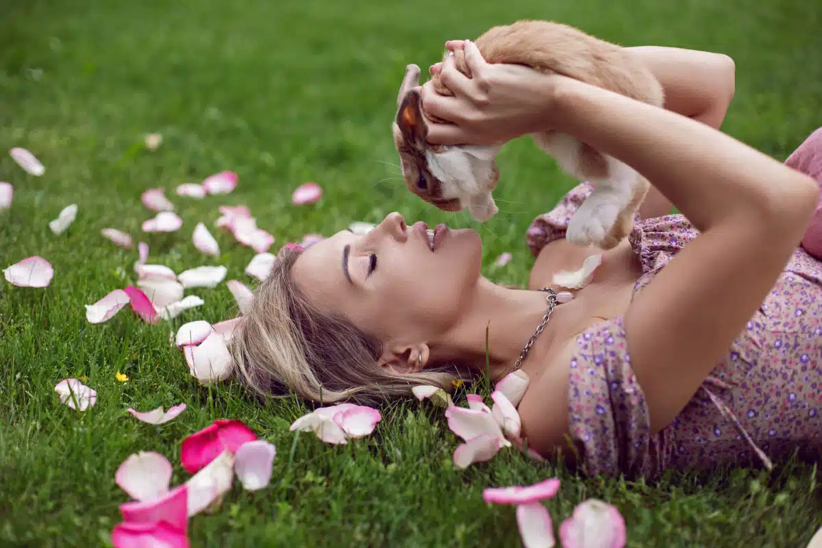 portrait of a beautiful young woman lying on a green meadow with rose petals and holding a rabbit in her hands in front of her face.