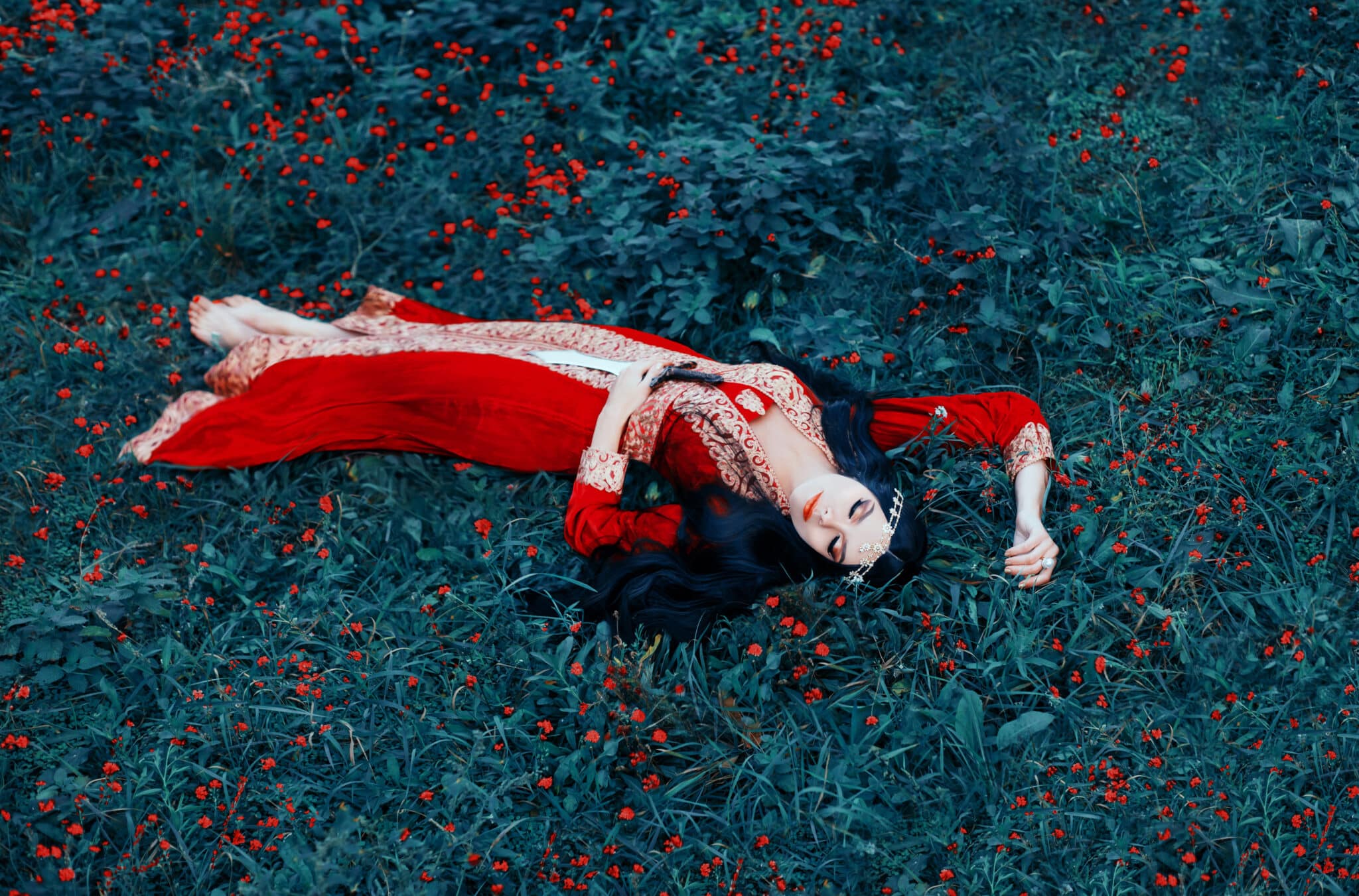 A young woman, a sleeping beauty, lies in a meadow of flowers in a chic red velvet dress, which is embroidered in gold with a sword in her hands. 