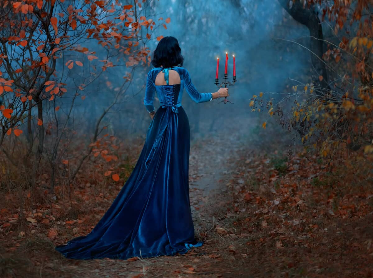 Silhouette fantasy woman queen runs in dark forest, hands hold vintage burning candlestick candles. Blue velvet long medieval dress. Gothic girl vampire princess back rear view. Autumn nature, trees