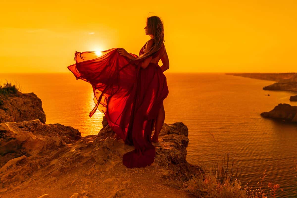 woman in red dress standing on a cliff with a beautiful view of the sunset and ocean