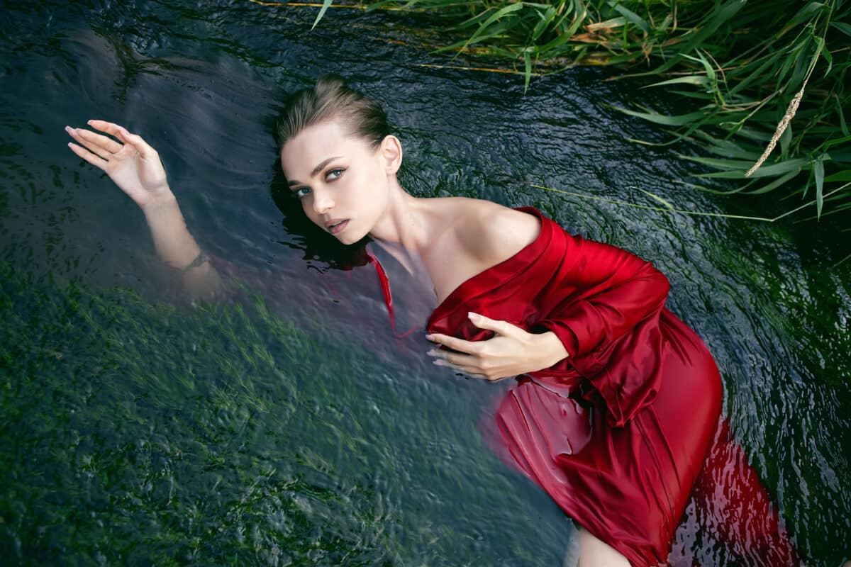 a romantic young woman in a red dress lying in a river with green algae in summer in nature during the day