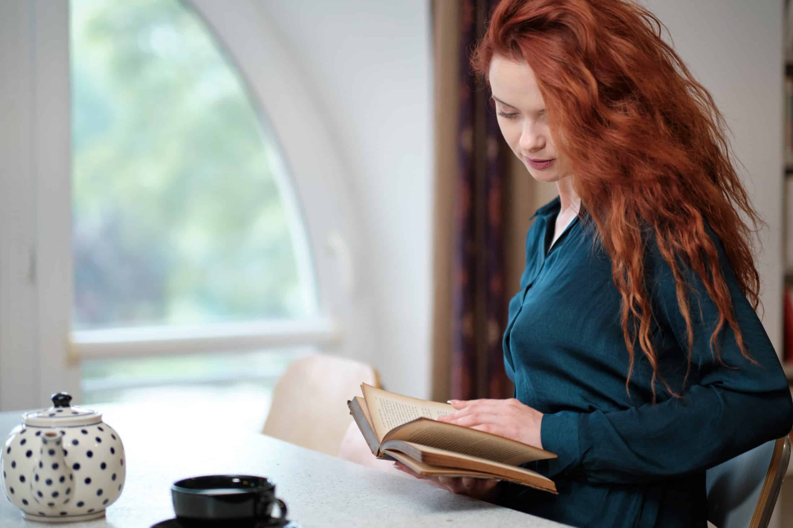 redhead woman reading book with coffee by the window
