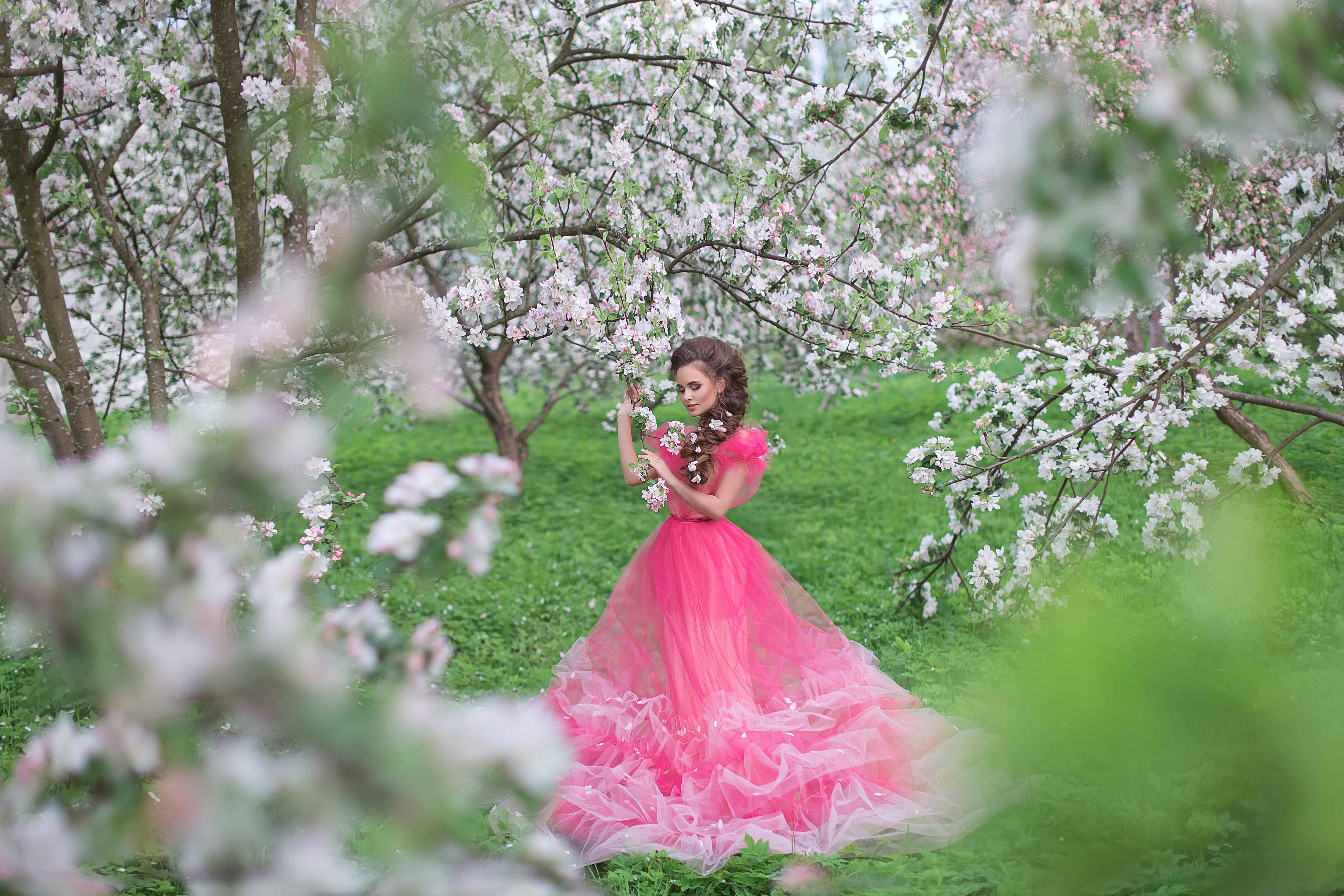 A beautiful young lady with long hair in a light pink ball gown in blooming orchard
