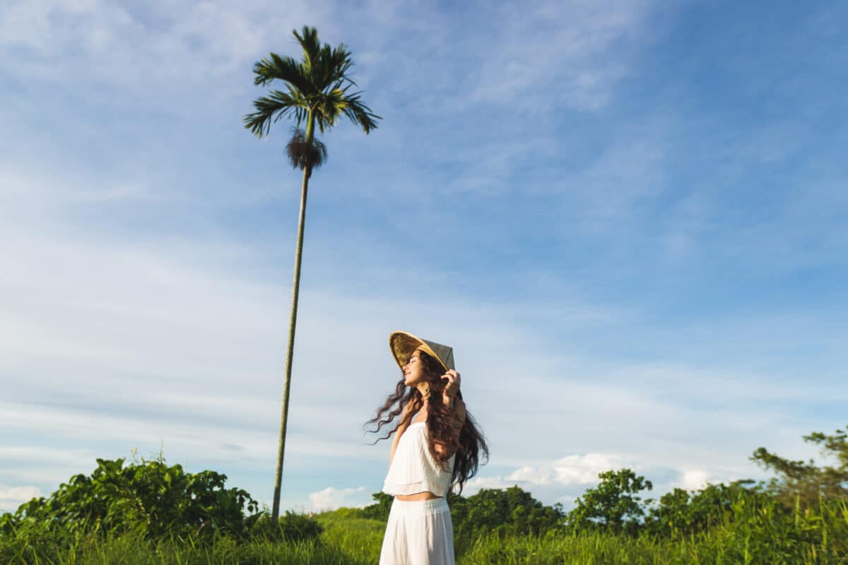 a young happy woman in a white traditional clothing standing in nature with a tall lone palm tree