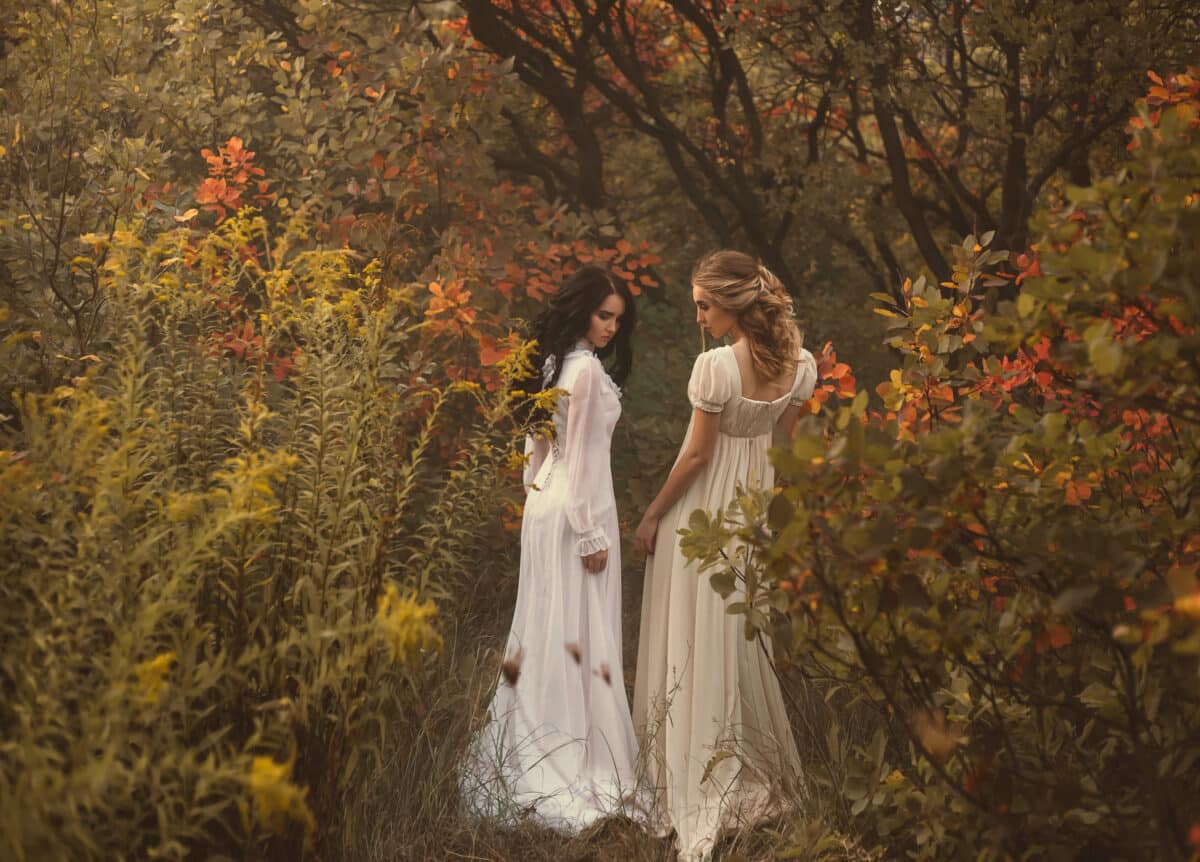 Two beautiful fairy sisters are walking in white vintage dresses in the woods