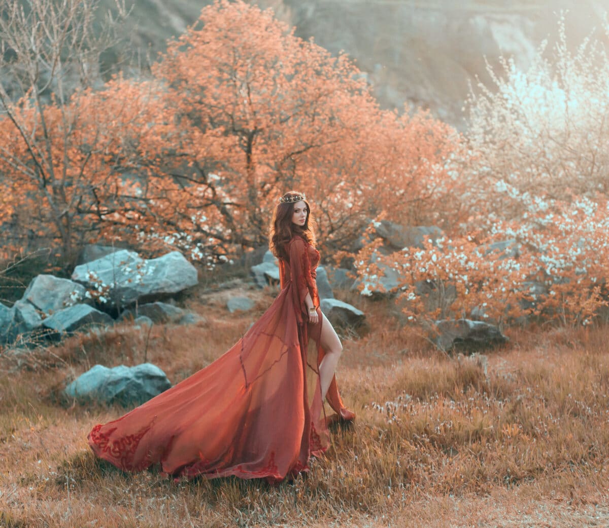A brunette girl with a golden crown and in a red dress in a long train walking at sunset. Background wildlife, flowering trees, sunlight, lush grass. Artistic photo