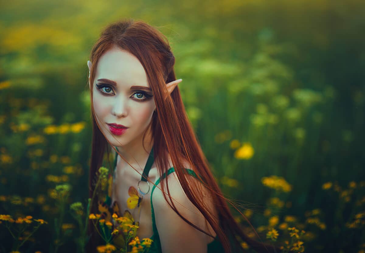 Portrait of a redheaded girl elf in a green swimsuit posing in a clearing of yellow flowers. Fantastic young woman with yellow butterflies in the rays of light.