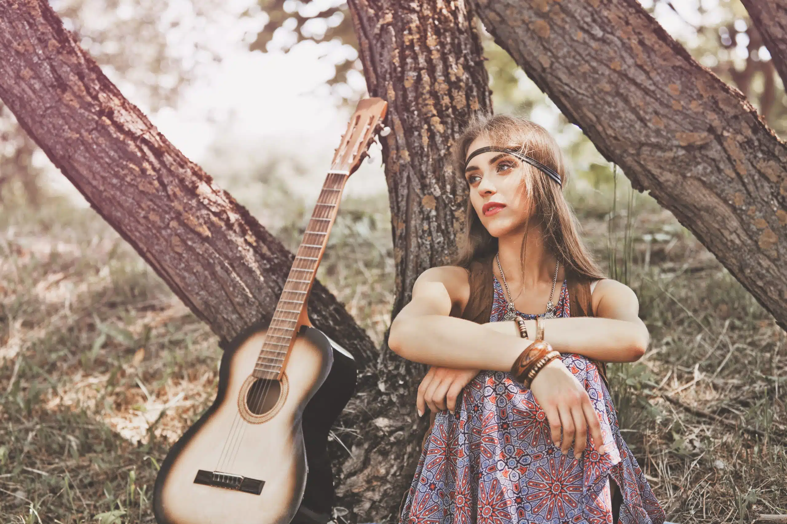 Pensive young hippie woman sitting near a tree, a guitar beside her