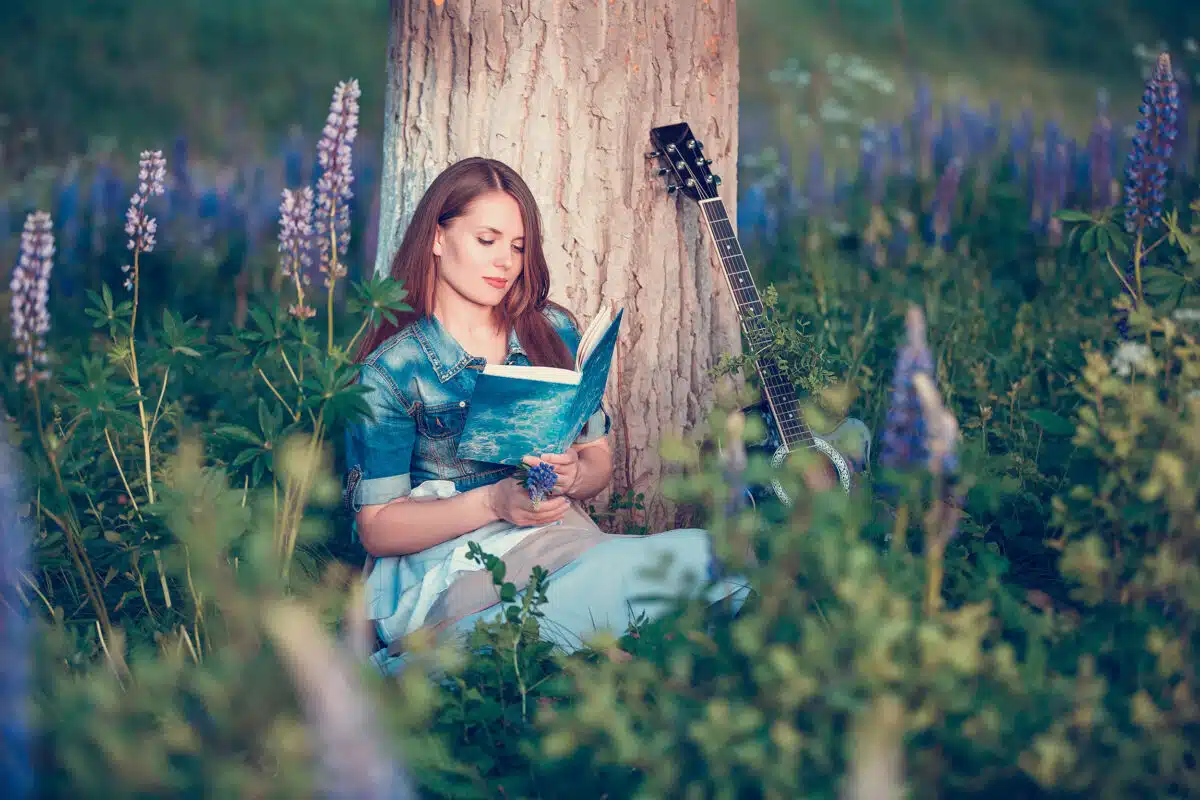 Beautiful woman near the tree and the field with lupine reads a book