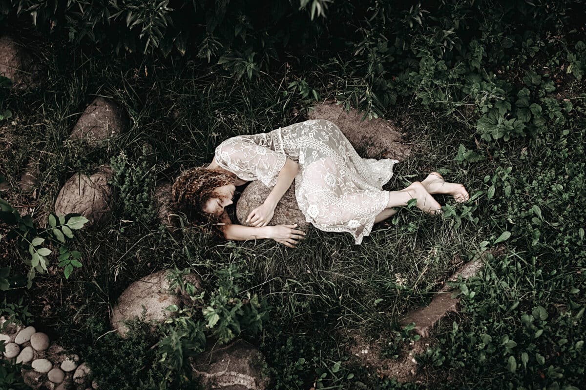 Young lonely woman in a white lace dress lying on green grass hugging a rock