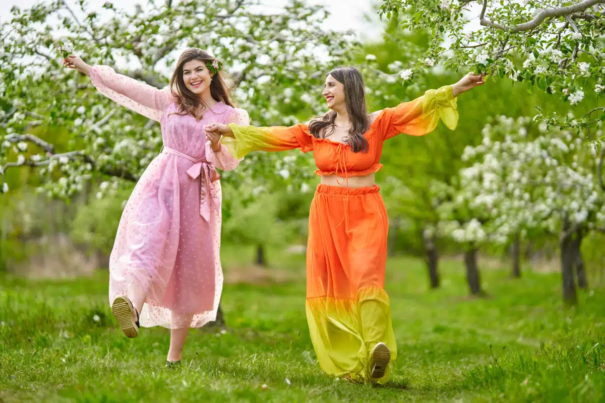 Two cheerful women walking hand in hand through an orchard