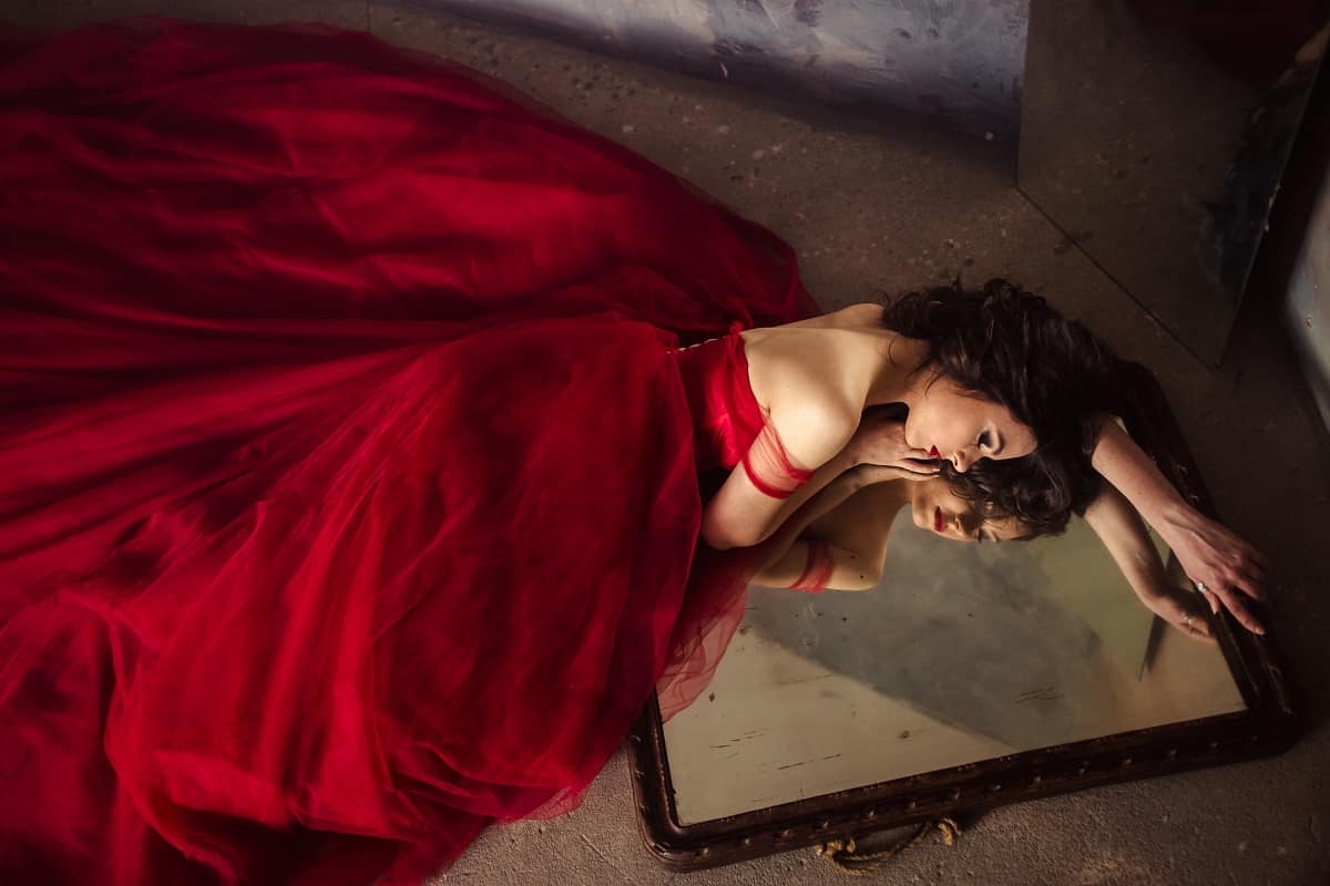 a sad but sensual woman in a long red dress lying on the ground on a mirror with her reflection